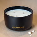 Black candle featuring the brand 'Aromavela' with a beige background and tiny broken seashells beside the candle.