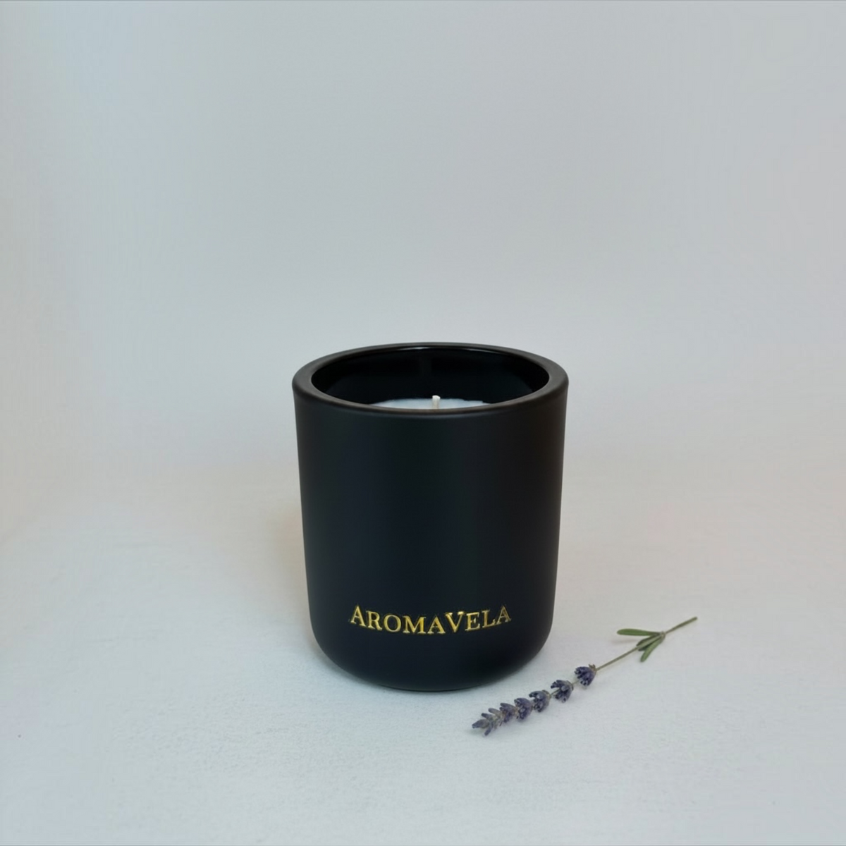 A matte black small cylindrical candle jar with gold 'AROMAVELA' lettering, shown on a light gray background with a small sprig of lavender beside it.